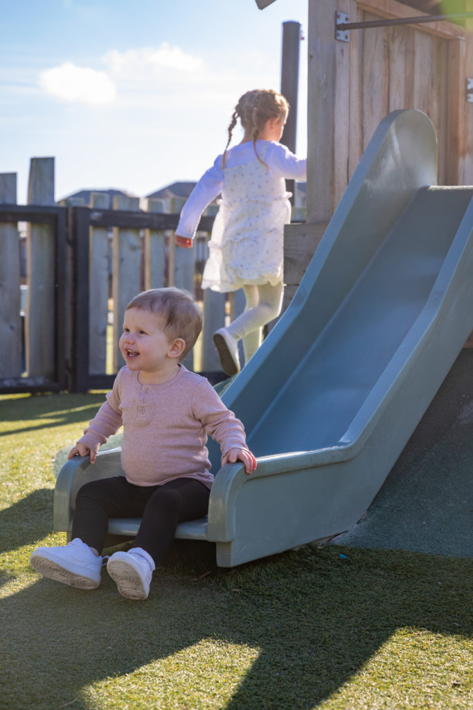 Smiling toddler at the bottom of a slide in The Stables Montessori outdoor play area, with another preschool child climbing the play structure, representing early childhood education and preschool Christchurch.