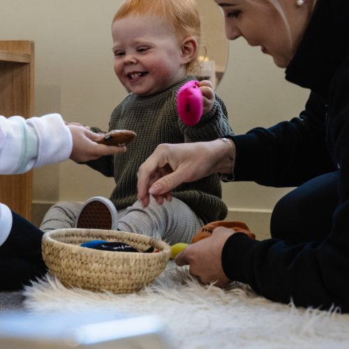 Smiling toddler exploring pretend food basket during Montessori early childhood activity, supported by teacher in preschool Christchurch learning environment.